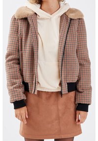 Röd och svart rutmönstrad bomberjacka med plyschkrage, svarta ribbade manschetter och dragkedjestängning, kombinerad med en krämvit hoodie och brun kjol.