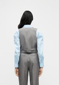 Γκρι προσαρμοσμένο γιλέκο με ζώνη, σε συνδυασμό με ανοιχτό μπλε πουκάμισο με μακριά μανίκια. Ομαλή υφή υφάσματος και καθαρές γραμμές σχεδίασης.