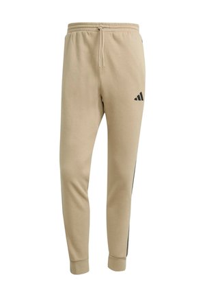 Pantaloni della tuta beige a gamba affusolata con vita elastica, coulisse, polsini a costine, logo Adidas nero sulla coscia superiore e dettagli a strisce laterali.