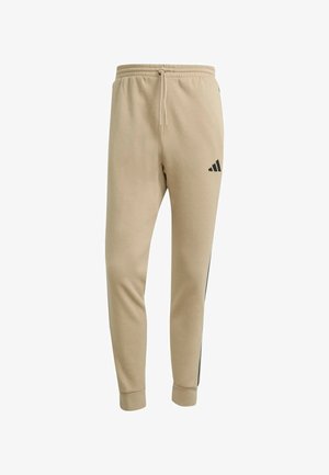Pantaloni della tuta beige a gamba affusolata con vita elastica, coulisse, polsini a costine, logo Adidas nero sulla coscia superiore e dettagli a strisce laterali.