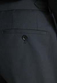 Schwarzer Stoff mit einer strukturierten Oberfläche, ausgestattet mit einer Tasche und einem einzelnen schwarzen Knopf an der Seite, was ein strukturiertes Design schafft.