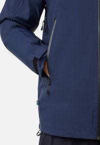Giacca impermeabile blu navy con una texture liscia, dotata di una tasca laterale con zip, fondo regolabile con cordoncino e un piccolo accento della bandiera svedese.