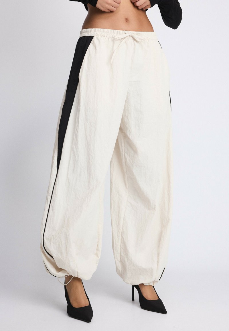 Pantalon blanc à jambes larges avec bandes latérales noires et poignets élastiques, porté avec des chaussures à talons noires et un top noir court.