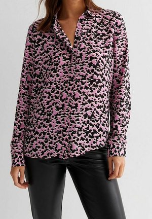 Kvinde iført langærmet pink bluse med sorte abstrakte pletter og sorte læderbukser, stående mod en ensartet lys baggrund.