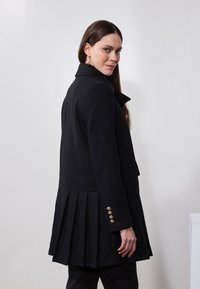 Manteau noir avec un design à dos plissé, en tissu doux ; dispose d'un col et de boutons dorés sur les manches.