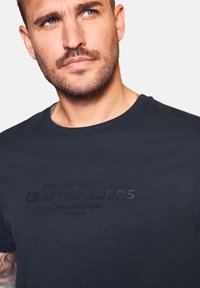 Muž s lehkým vousem a modrýma očima, oblečený v tmavě modrém tričku s embosovaným nápisem "Ruční zboží" na hrudi, dívající se mírně vzhůru.