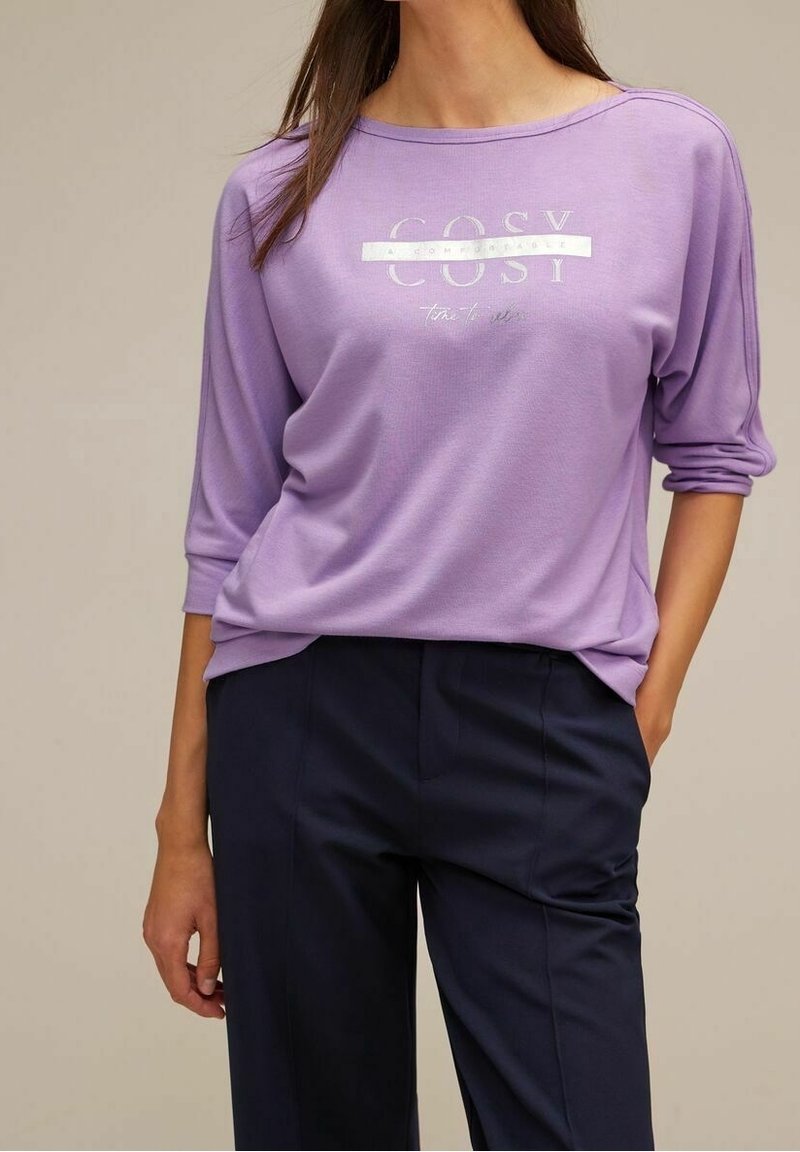 Ljust lila långärmad topp med en bred nacke, prydd med en silverfärgad textgrafik som skriver "COSY" och en avslappnad passform, i kombination med mörkblå byxor.