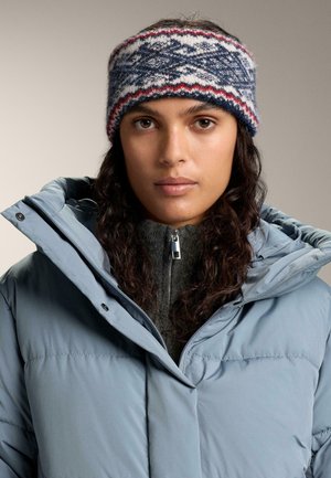 Fascia per capelli lavorata a maglia con motivo blu, bianco e rosso. Presenta una texture morbida. Indossata con una giacca imbottita azzurro chiaro e un maglione grigio con zip.
