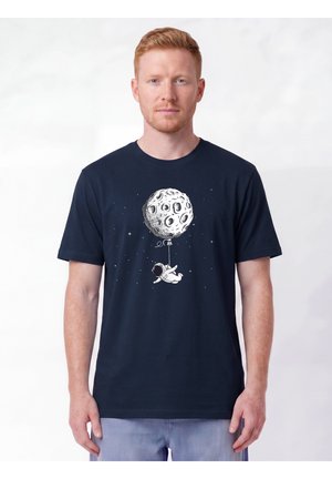 Homme portant une chemise bleu foncé avec un motif d'astronaute flottant dans l'espace tenant une ficelle attachée à un grand ballon en forme de lune.