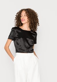 Top cropped em satin preto com mangas curtas, decote redondo, textura suave, combinado com calças brancas de cintura alta, mãos nos bolsos.