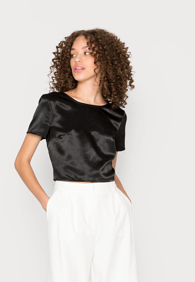 Top cropped em satin preto com mangas curtas, decote redondo, textura suave, combinado com calças brancas de cintura alta, mãos nos bolsos.