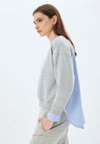 Sudadera gris con un panel trasero a rayas azules, que cuenta con puños acanalados y un ajuste relajado. Tela suave con texturas y colores contrastantes.