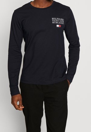 Man wearing a navy long-sleeve shirt with "Hilfiger Yacht Club MCMLXXXV" text and a small red, white, and navy flag on chest.