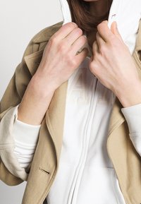 Beige jacket over a white zip-up hoodie. The jacket has a structured design with visible stitching, while the hoodie features a subtle logo.