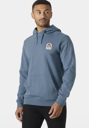 Hombre con sudadera azul con capucha y bolsillo delantero, con pequeño logo de temática natural en el pecho izquierdo, mirando hacia su izquierda.