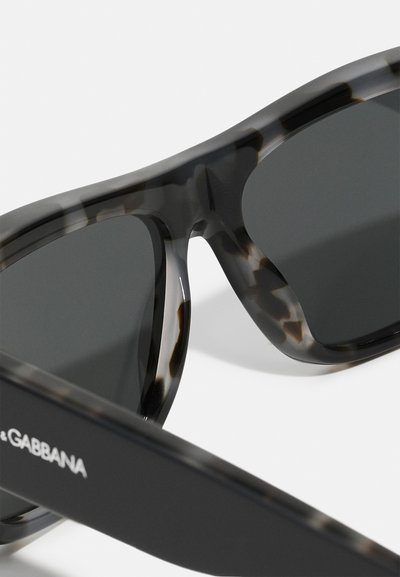 Ochelari de soare din tortoiseshell închise, cu un design elegant și curbat. Prezintă lentile întunecate și un braț mat negru cu detaliu logo.