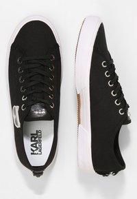 Musta matalavartinen tennari, jossa on valkoiset kumipohjat, mustat nauhat, metalliset nauhakutdaterit ja "Karl Lagerfeld" -merkintä sisäpohjassa ja kantapäässä.