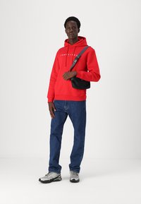 Raudona gobtuvė su "TOMMY JEANS" logotipu, derinama su mėlynais džinsais ir pilkais sportbačiais. Juoda petinė kuprinė per petį.