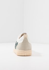 Białe sneakersy z zielonymi zamszowymi akcentami, teksturowanym beżowym obcasem i gumową podeszwą. Minimalistyczny design z gładkimi i miękkimi materiałami.