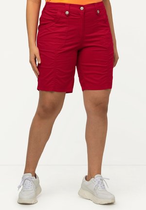 Personne portant un short rouge mi-long avec des poches avant et des baskets blanches, debout devant un fond clair uni.