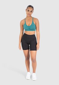 Teal urheilu-rintaliivit, joissa on rypytys edessä, yhdistettyinä korkea vyötärö black shorts -shortseihin. Malli seisoo valkoisissa sneaksereissä vaaleataustalla.