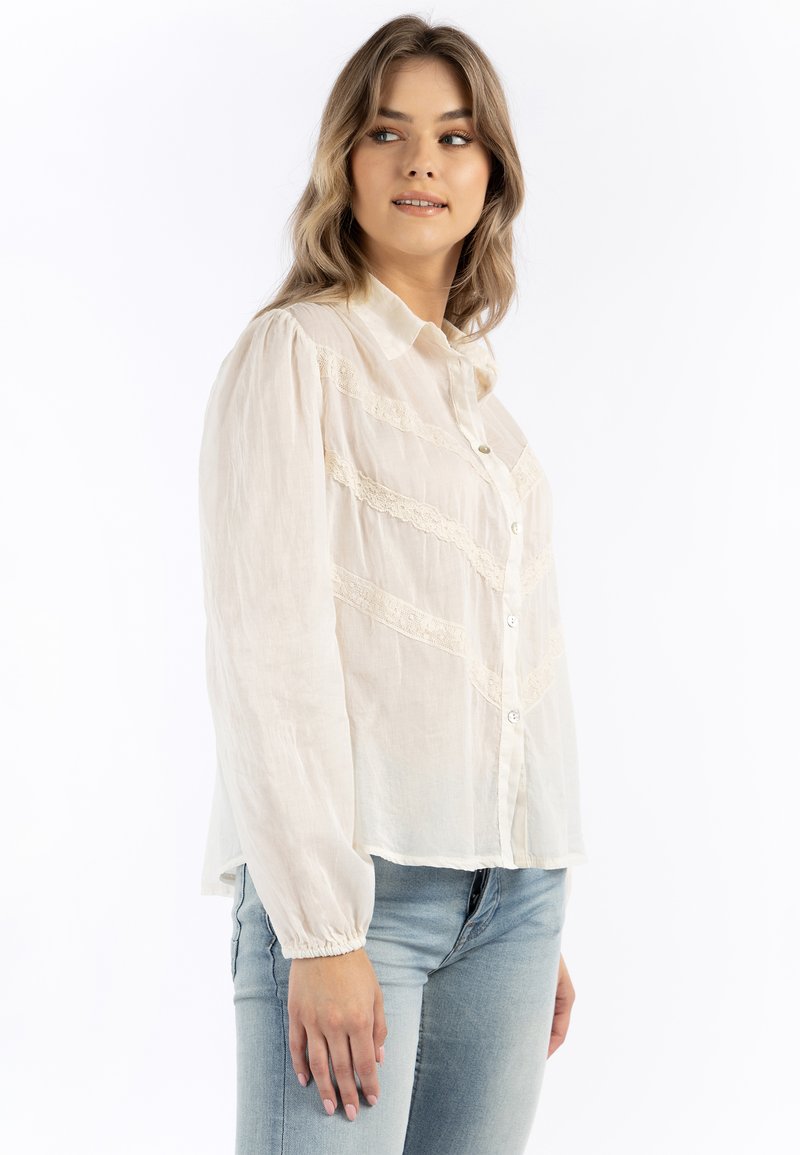 Blusa beige chiaro con chiusura a bottoni, dettagli in pizzo e maniche lunghe con polsini elastici. Indossata con jeans azzurri.