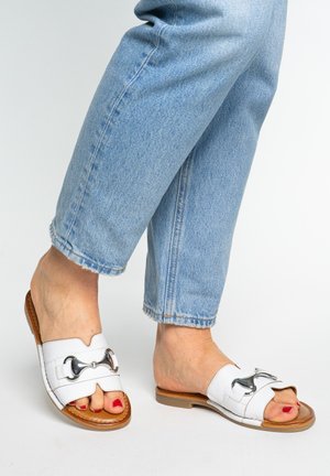 Pieds portant des sandales en cuir blanches à enfiler avec des détails de boucle argentée, associées à un jean court bleu clair et un vernis à ongles rouge.