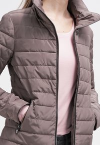 Puchowa kurtka w stonowanym taupe z wysokim kołnierzem i zapięciem na zamek. Posiada boczne kieszenie oraz pikowany wzór dla dodatkowej tekstury.