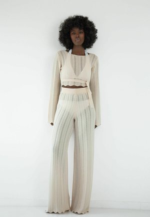 Spleciony zestaw dwuczęściowy w jasnym beżu, składający się z krótkiego swetra z długim rękawem oraz szerokich spodni z wzorem w jodełkę i prążkowaną fakturą.