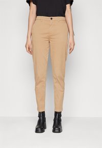 Personne portant un pantalon tapered beige, un haut noir à manches courtes et des bottines à talons noires, se tenant devant un fond gris uni.