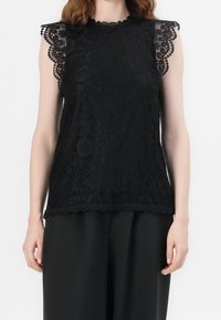 Top in pizzo nero con maniche e orlo a scallop. Presenta motivi intricati e una finitura testurizzata, indossato con pantaloni neri larghi.
