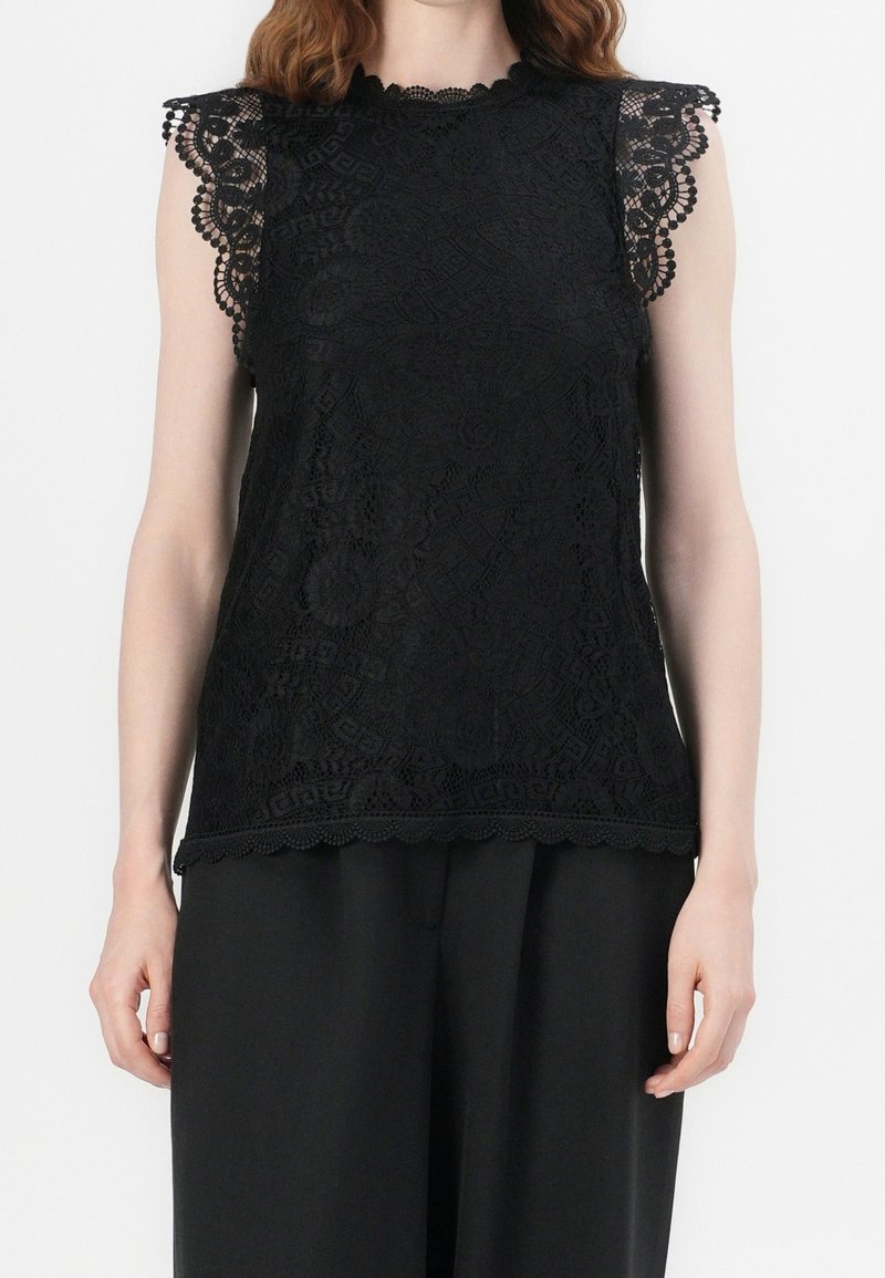 Top in pizzo nero con maniche e orlo a scallop. Presenta motivi intricati e una finitura testurizzata, indossato con pantaloni neri larghi.