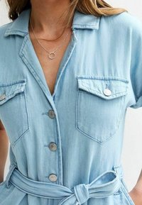 Vaaleansininen farkkumekko, jossa on kaulus, lyhyet hihat, kaksi rintataskua, napitettava etuosa ja kangasvyö. Keskiverto rakenne ja hillitty ompelu.