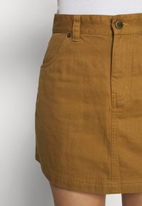 Mini-jupe en denim marron avec poches à l'avant, passants pour ceinture et fermeture par boutons, portée avec un haut blanc.