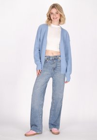 Cardigan lavorato a maglia blu chiaro con maniche lunghe sopra un crop top bianco, abbinato a jeans a gamba larga blu e scarpe rosa chiaro.