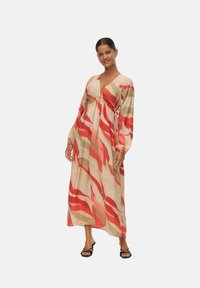 Femme portant une robe longue et ample avec des motifs abstraits rouges, beige et bruns, posant avec une main détendue, souriante à la caméra.