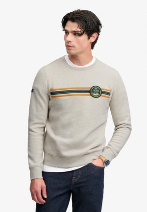 Mand iført en lysebeige sweatshirt med horisontale grønne og orange striber og et rundt mærke, kombineret med mørke jeans og et armbåndsur.
