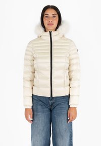 Giacca puffer beige con zip frontale, polsini a costine e un morbido cappuccio in pelliccia sintetica. Presenta pannelli cuciti per l'isolamento. Abbinata a jeans blu.