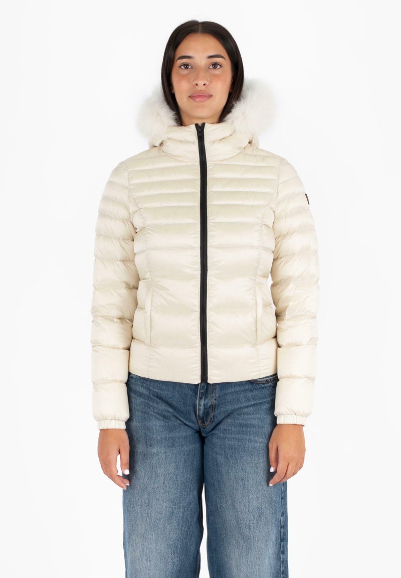 Giacca puffer beige con zip frontale, polsini a costine e un morbido cappuccio in pelliccia sintetica. Presenta pannelli cuciti per l'isolamento. Abbinata a jeans blu.