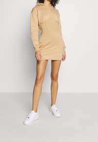 Vestido beige con capucha, con un cuerpo entallado y un corte relajado, combinado con zapatillas blancas. Tela suave, diseño de longitud a la rodilla y mangas largas.