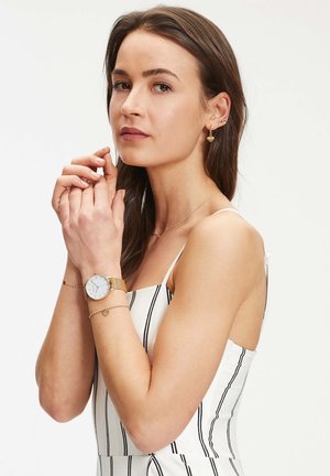 Goldfarbene Netzarmbanduhr mit einem weißen Zifferblatt, kombiniert mit goldenen, herzförmigen Ohrringen und zarten Armbändern, getragen zu einem gestreiften, ärmellosen Oberteil.