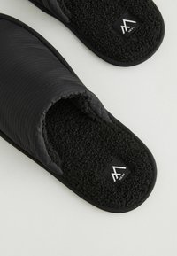 Chaussons noirs avec un extérieur matelassé, doublure en polaire noire douce et semelle texturée. Comprend une étiquette logo près du talon.