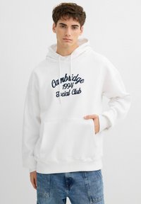 Sudadera con capucha blanca con texto bordado en color azul marino "Cambridge 1994 Social Club", bolsillo canguro, capucha con cordón y corte relajado. Textura de tela ligera.