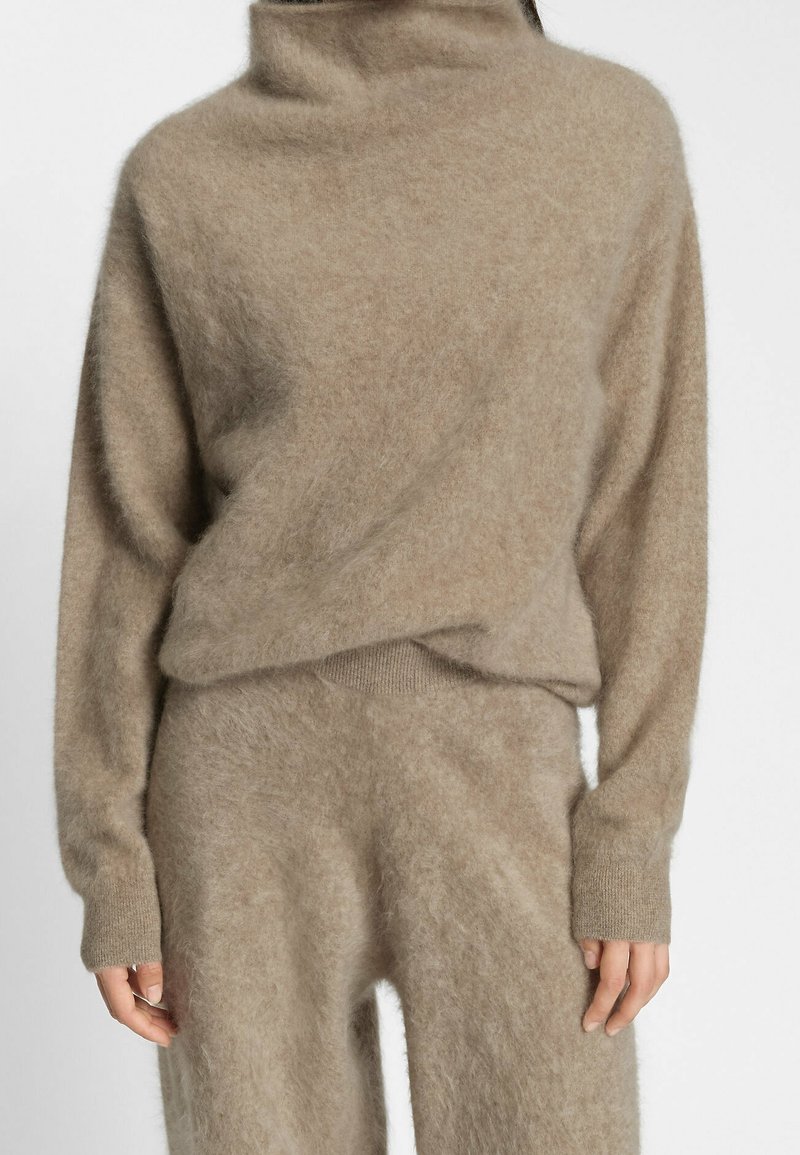 Personne portant un pull à col roulé beige, ample et duveteux, avec un pantalon assorti, sur fond blanc uni. Haut du corps visible.