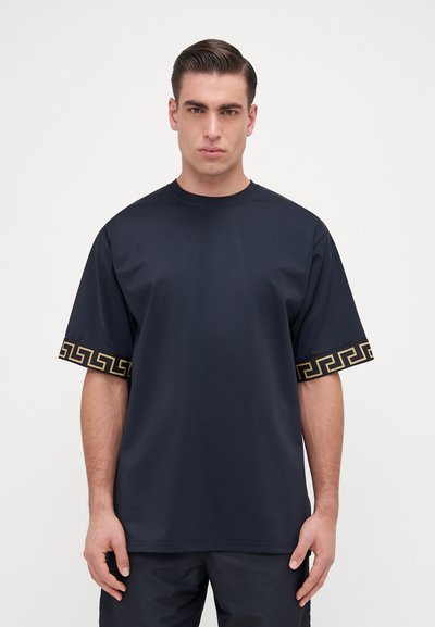 T-shirt oversize blu navy con maniche corte, caratterizzato da un motivo geometrico dorato lungo i bordi delle maniche. Tessuto morbido, design con collo a giro.