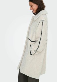 Cappotto di colore crema, soffice e peloso, con una superficie testurizzata, caratterizzato da accenti neri e tasche. Il design include un grande colletto e spacchi laterali.