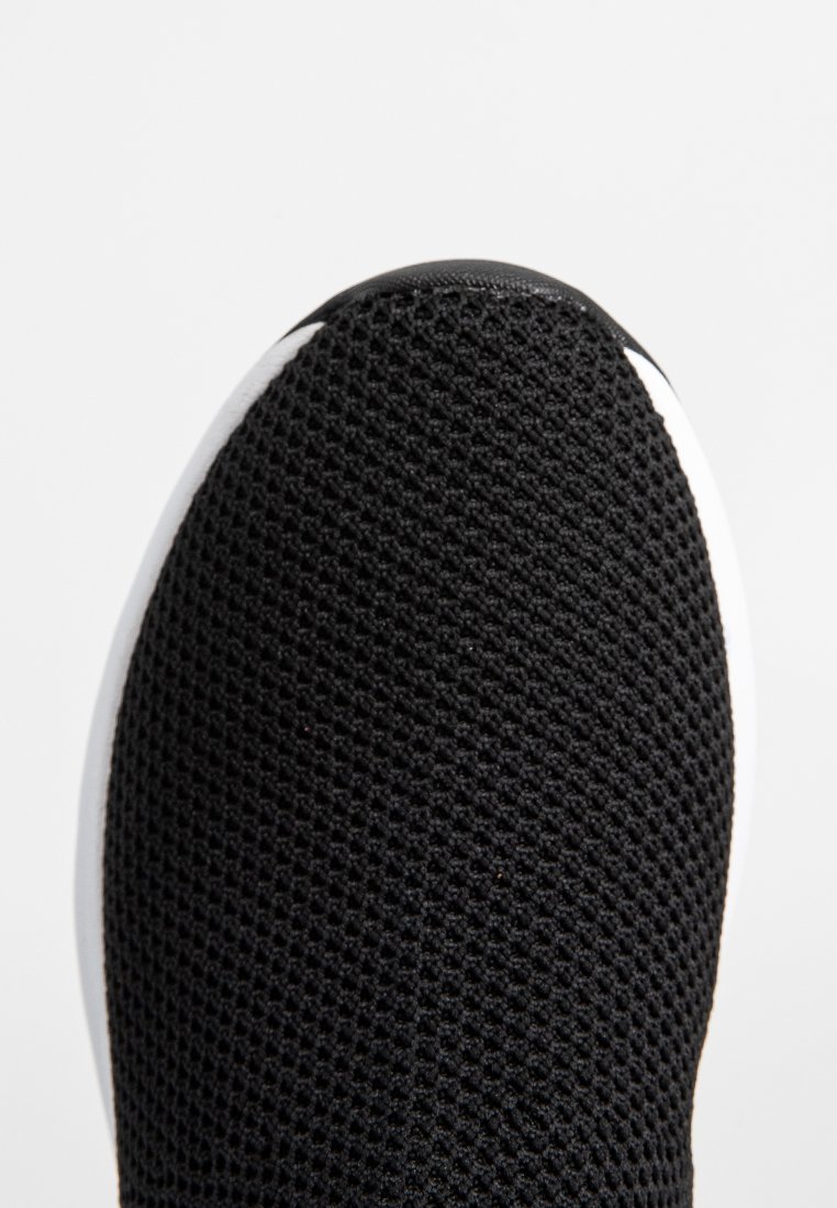 Gros plan sur l'embout d'une chaussure en tissu maillé noir avec un bord de semelle blanc, sur un fond blanc uni.