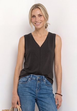 Smilende kvinde med blond hår trukket tilbage, iført en sort ærmeløs V-hals top og blå jeans, der holder i håndtaget på en flettet taske.