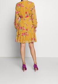 Blommig klänning i senapsgul med rosa och gröna accenter, med långa ärmar, utsvängd nederkant och ett matchande bälte. Sammanställd med lila högklackade skor.