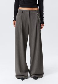 Femme portant un pantalon gris à rayures verticales, taille haute et jambes larges, avec un accessoire en chaîne argentée, associé à un haut noir à manches longues cropped.